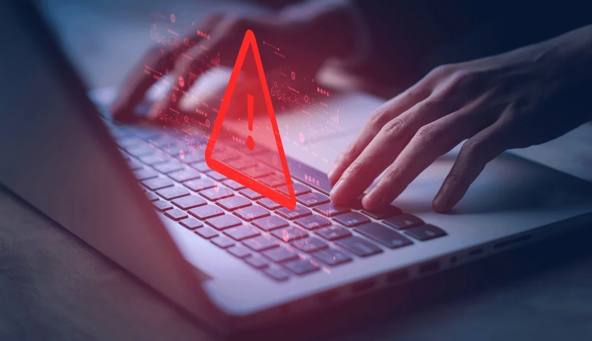 A person's hands typing on a laptop, overlaid with a red caution symbol, indicating a cybersecurity alert or warning.