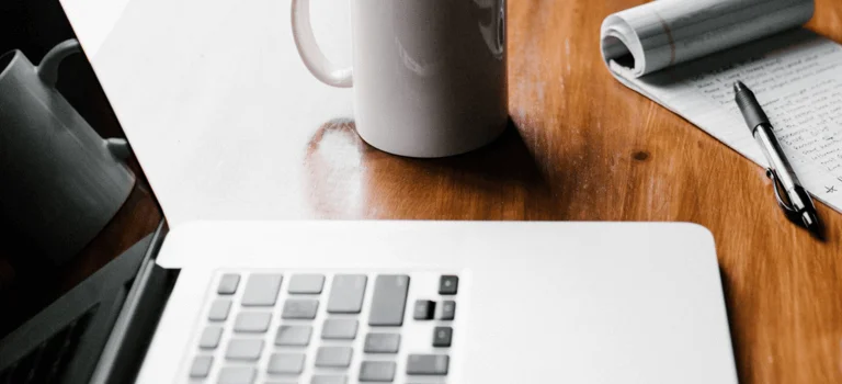 A wooden desk with a laptop, coffee mug, notepad, pen, and smartphone, creating a cozy workspace atmosphere.