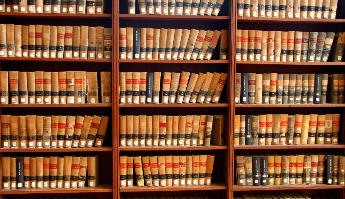 Rows of aged books neatly arranged on wooden shelves, featuring red and black spines with labels, in a historic library setting.