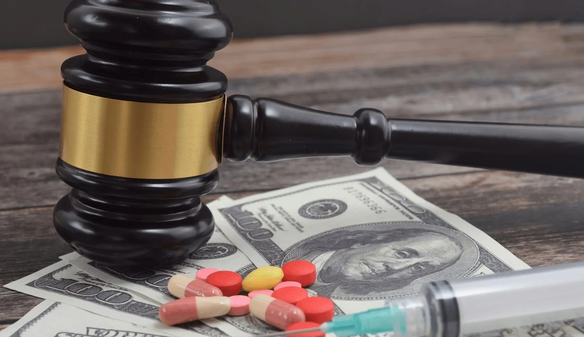 A gavel rests on money, accompanied by various pills and a syringe, symbolizing legality and the pharmaceutical industry.