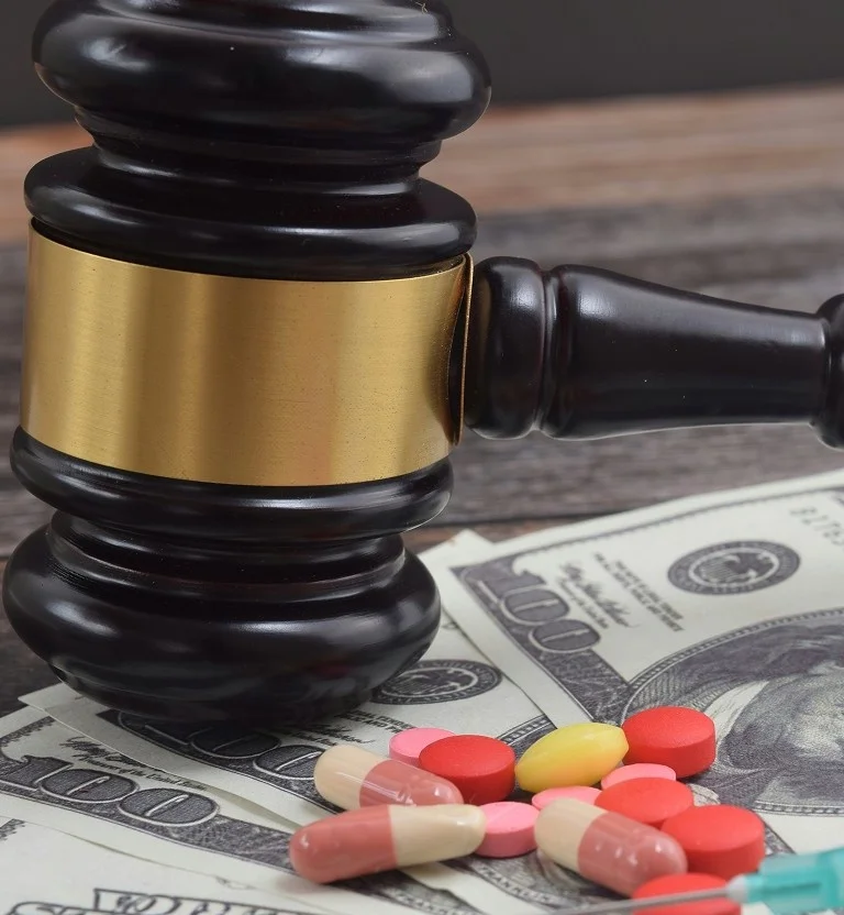 A gavel rests on money, accompanied by various pills and a syringe, symbolizing legality and the pharmaceutical industry.