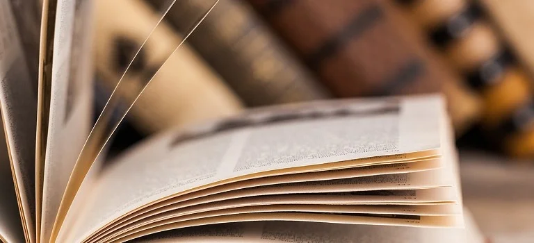 An open book resting on a table, with blurred vintage books in the background and a pair of reading glasses nearby.
